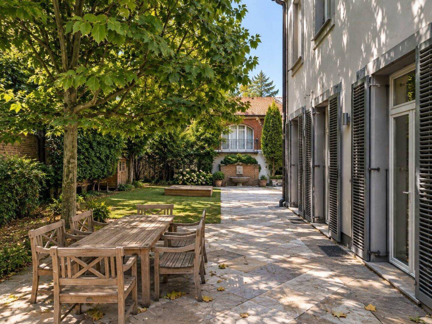 Große Steinterrasse mit Holzesstisch und Stühlen, umgeben von einem gepflegten Garten und Bäumen.