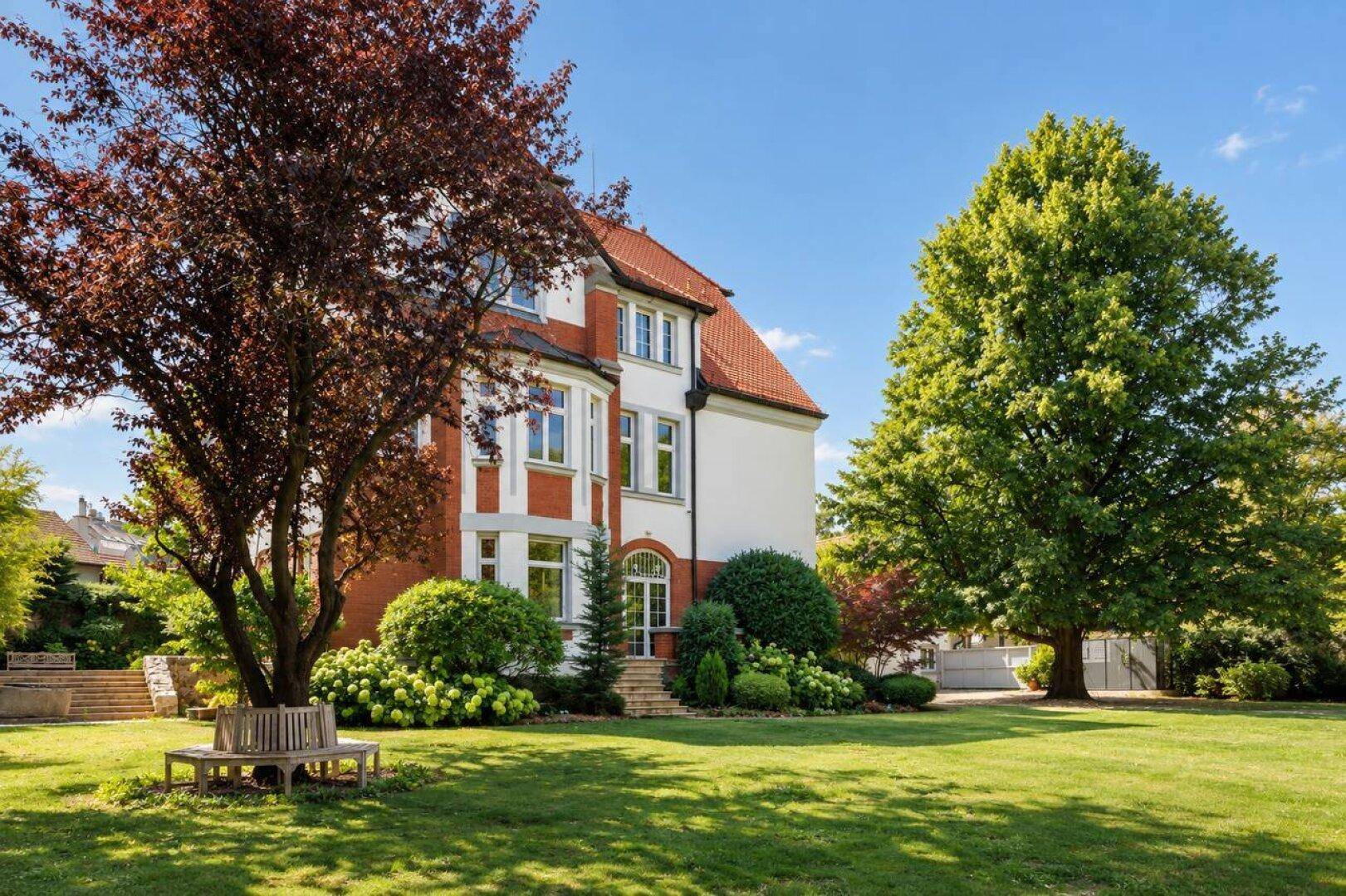 Historische Villa mit rotem Ziegeldach, gepflegtem Rasen und schattenspendenden Bäumen an einem sonnigen Tag.