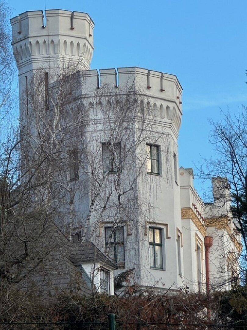 Detailansicht des historischen Turms mit Zinnen und Fenstern, umgeben von kahlen Bäumen.