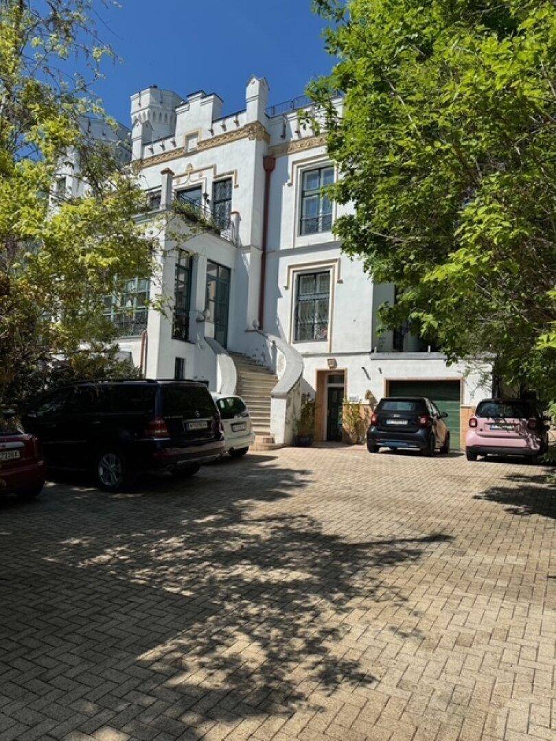 Eingangsbereich der historischen Villa mit gepflasterter Auffahrt und parkenden Fahrzeugen im Sommer.