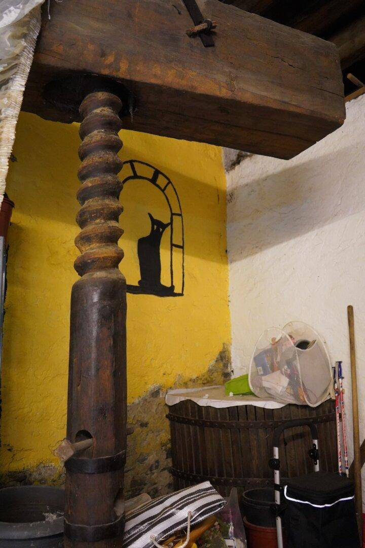 Kellerraum mit einer alten Holzpresse, gelben Wänden und sichtbaren Holzbalken.