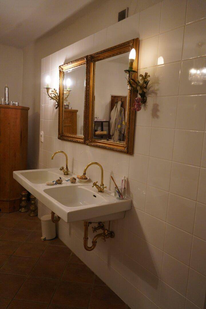 Badezimmer mit Doppelwaschbecken, goldenen Armaturen und einem großen Spiegel mit Wandleuchten.
