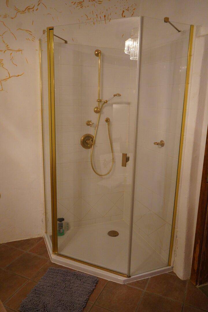 Badezimmer mit einer Eckdusche und goldenen Armaturen, gefliestem Boden und Wänden.