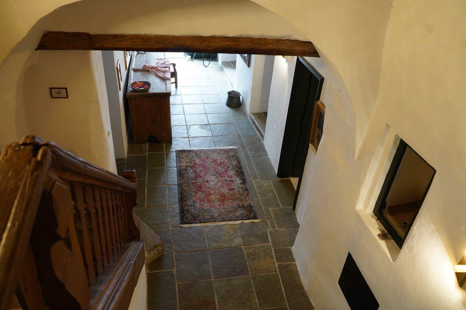 Blick von der Holztreppe in einen Flur mit Steinboden und rustikalen Holzbalken an der Decke.