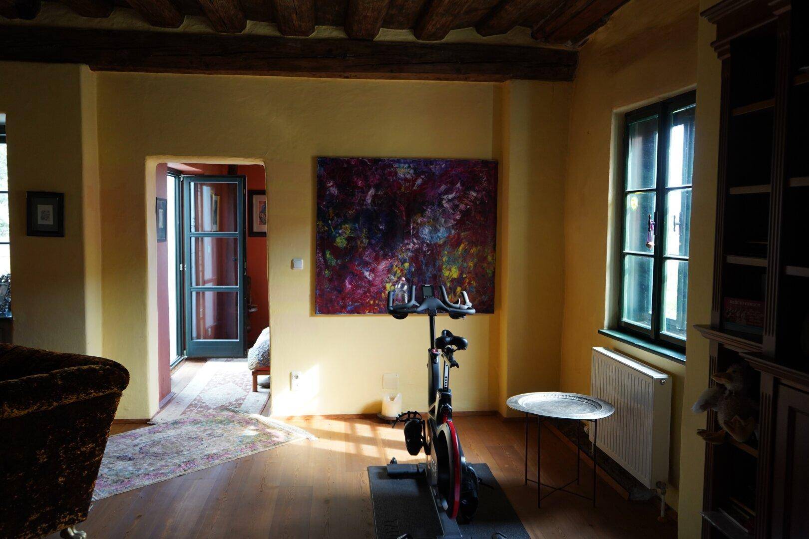 Heller Wohnbereich mit Trainingsrad, Fenster und Zugang zu einem weiteren Zimmer.