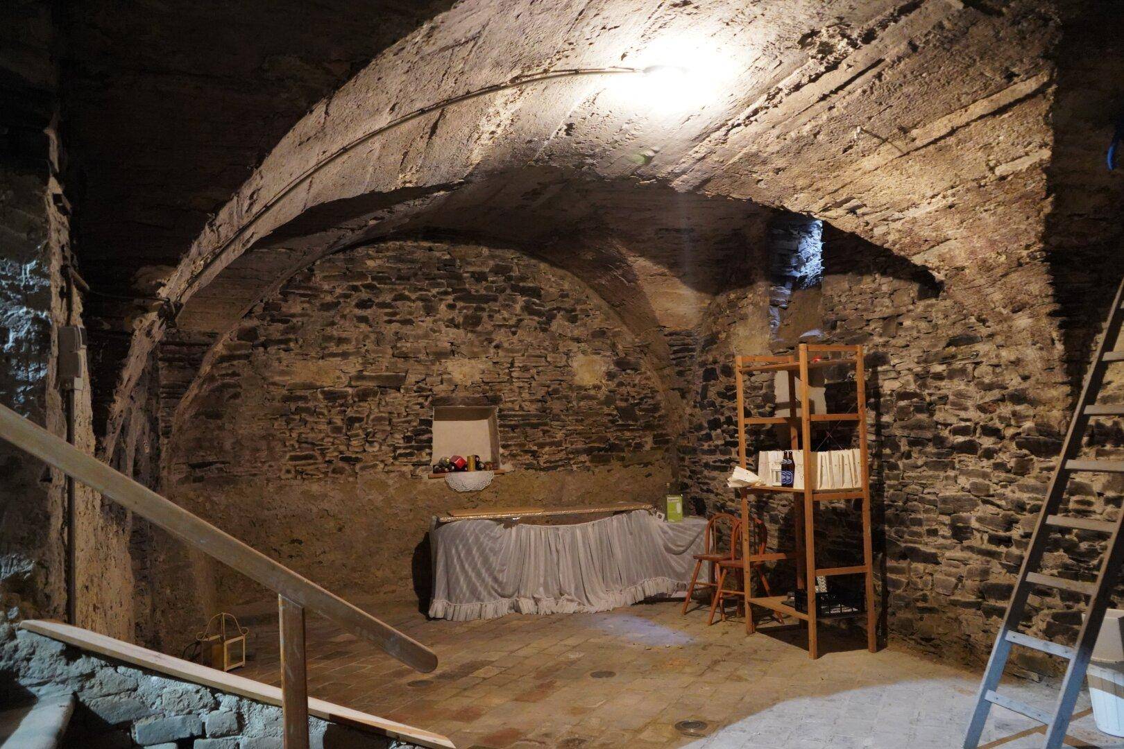 Rustikaler Gewölbekeller mit Steinmauern, Holzbalken und einfacher Einrichtung, beleuchtet von einer einzelnen Lampe.