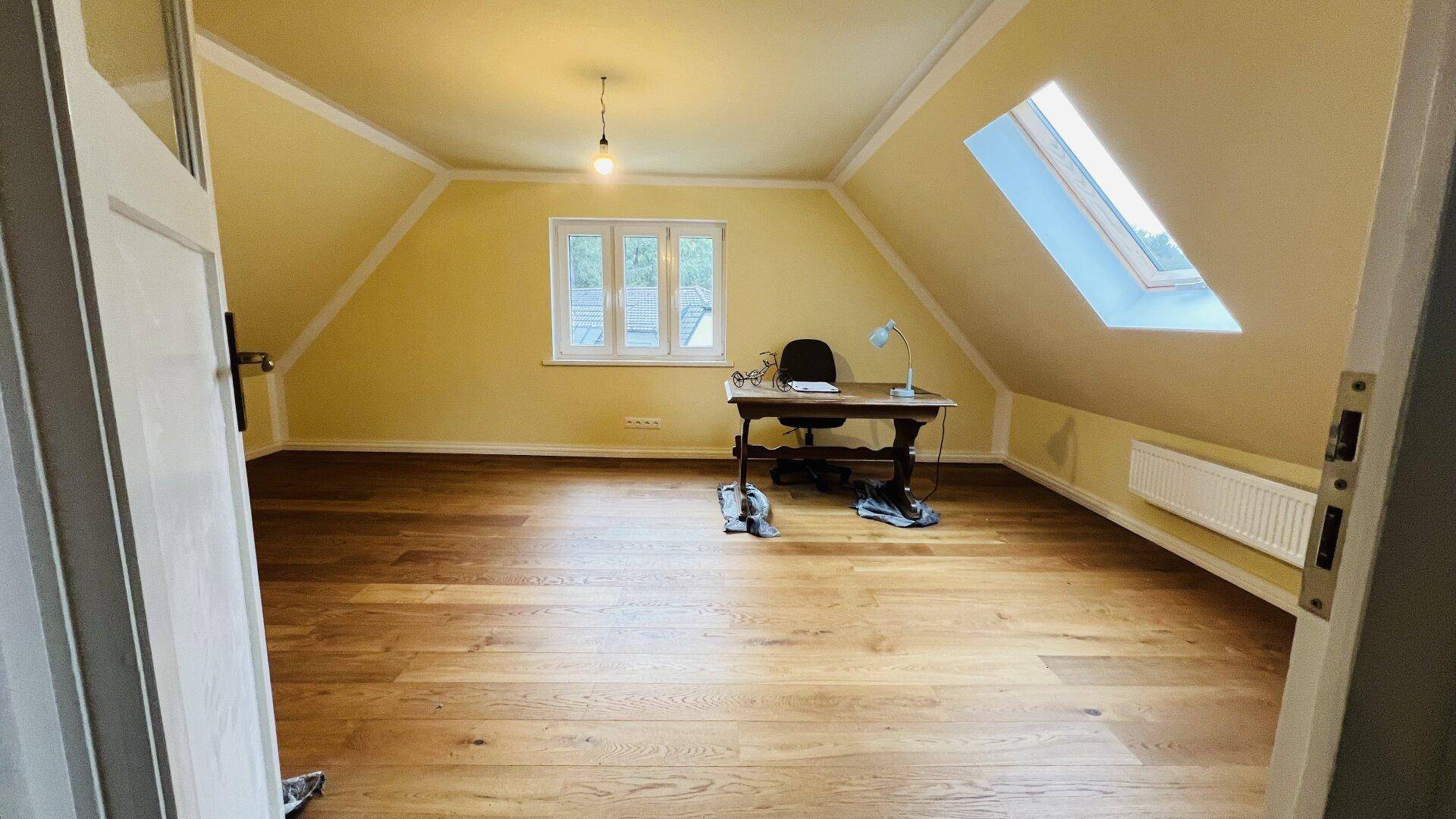 Helles Dachzimmer mit Holzboden, Dachschräge und einem Arbeitsbereich am Fenster.