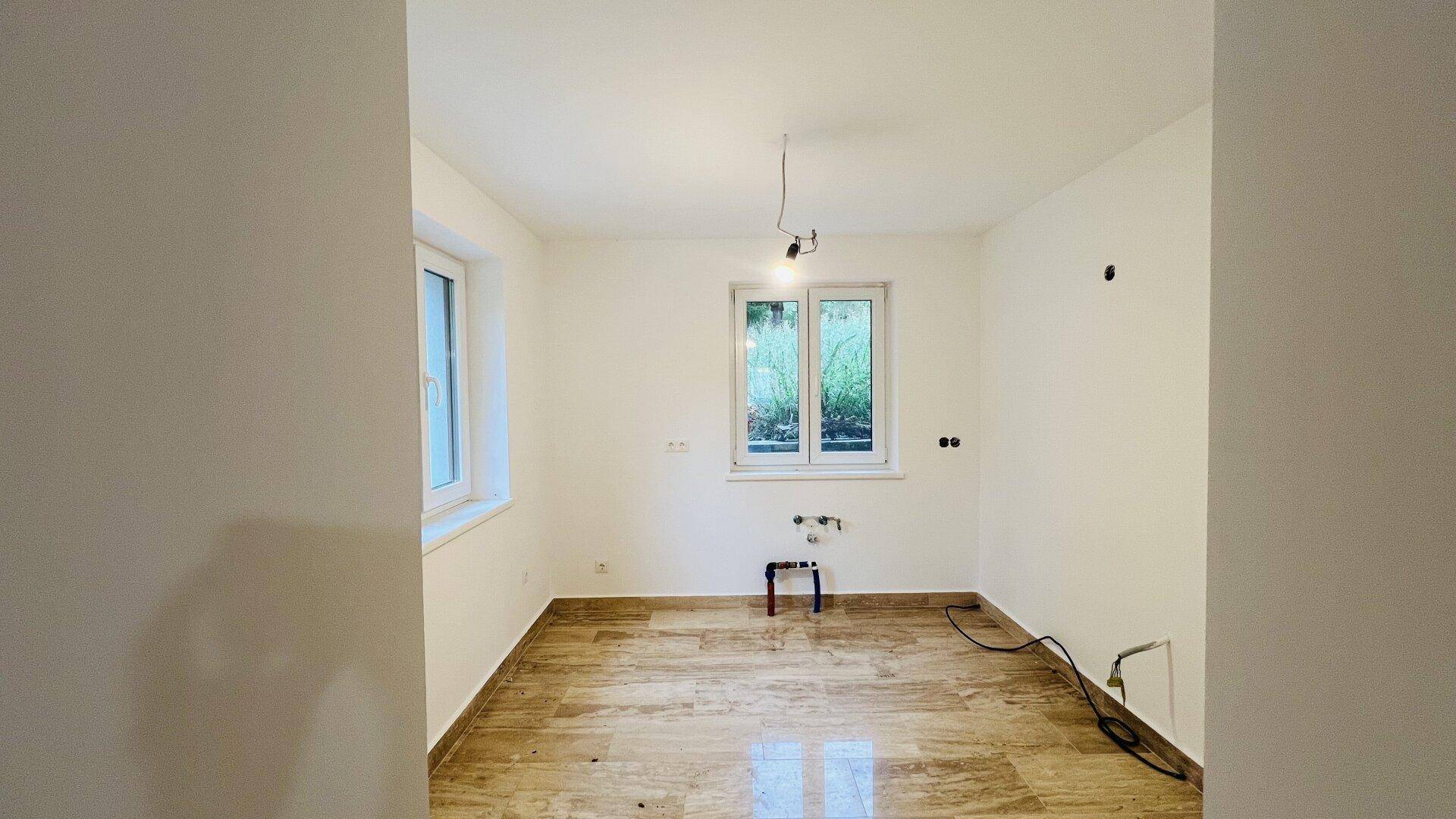 Leerer Raum, vorbereitet für eine Küche, mit sichtbaren Wasseranschlüssen und Fliesenboden.