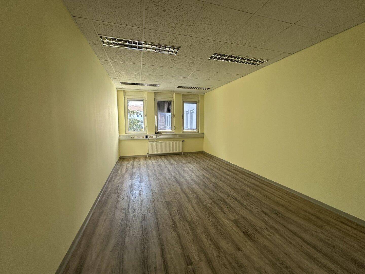 Helles, leeres Büro mit Laminatboden und Fenstern, die viel Tageslicht hereinlassen.