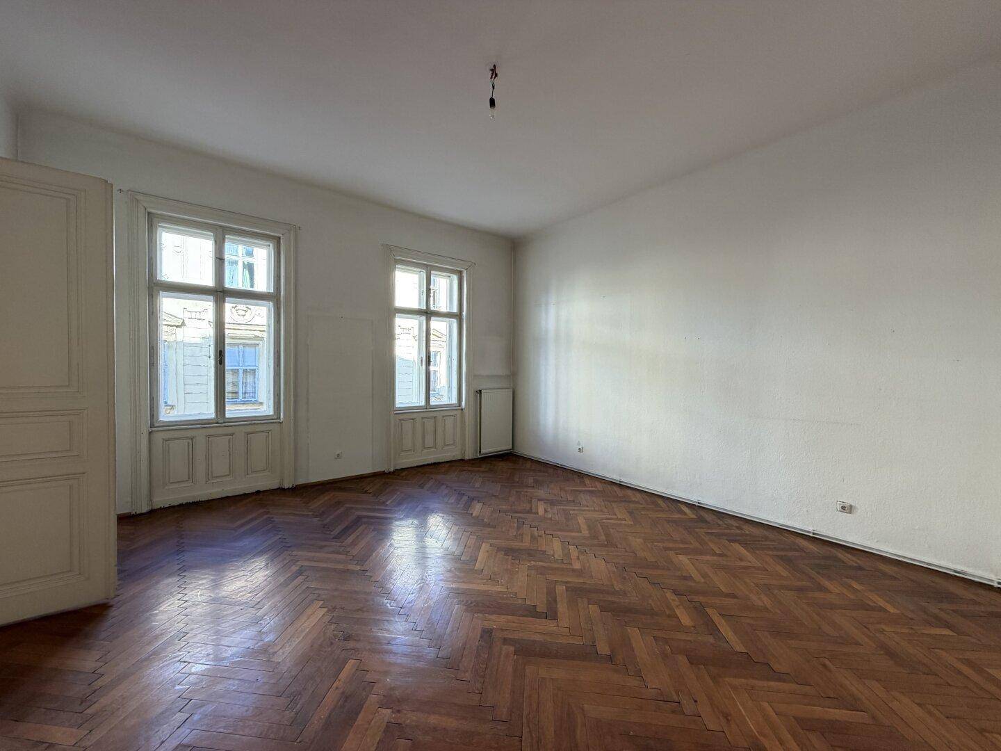 Helles Wohnzimmer mit Fischgrätparkett und zwei hohen Fenstern, die viel Licht spenden.