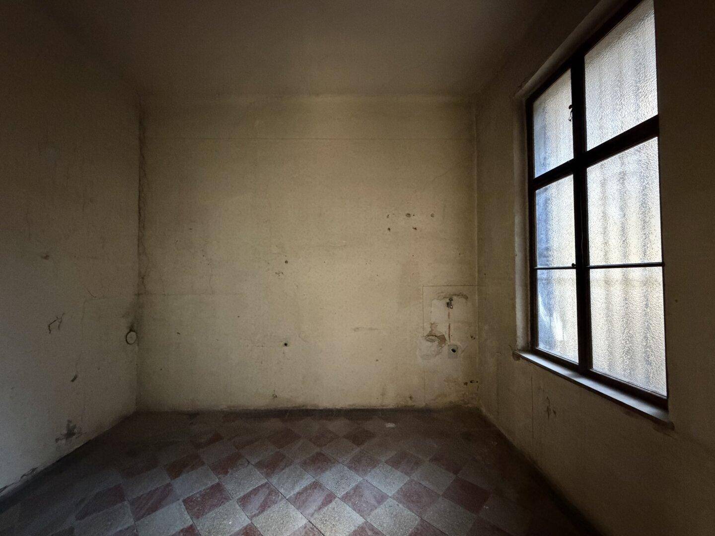 Kleiner Raum mit kariertem Fliesenboden und einem Fenster, das wenig Licht spendet.