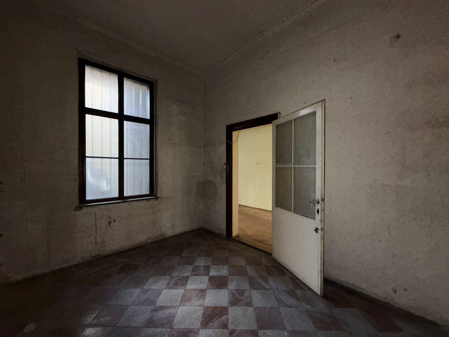 Dunkler Raum mit kariertem Fliesenboden und einer alten Holztür, die in einen weiteren Raum führt.