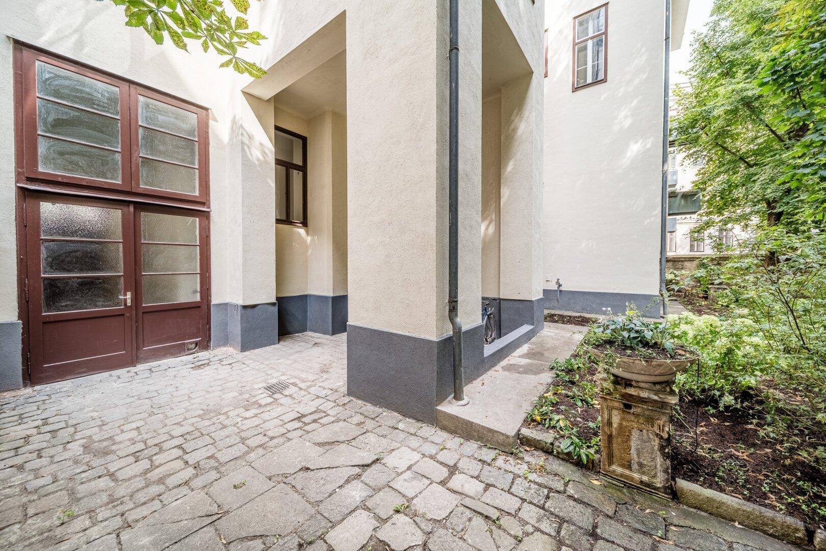 Gepflasterter Außenbereich neben dem Gebäude mit Zugang zu einem begrünten Garten.