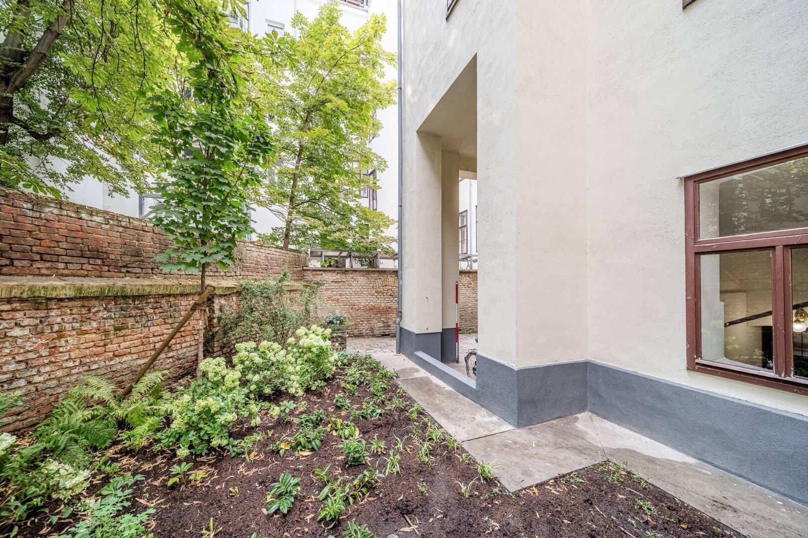 Ruhiger Gartenbereich mit üppigem Grün und einer alten Ziegelmauer im Hintergrund.