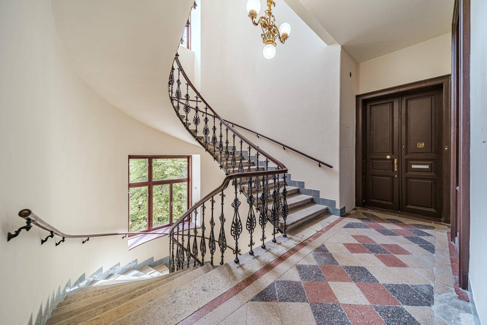 Elegantes Treppenhaus mit geschwungenem Geländer, hohen Decken und großen Fenstern.