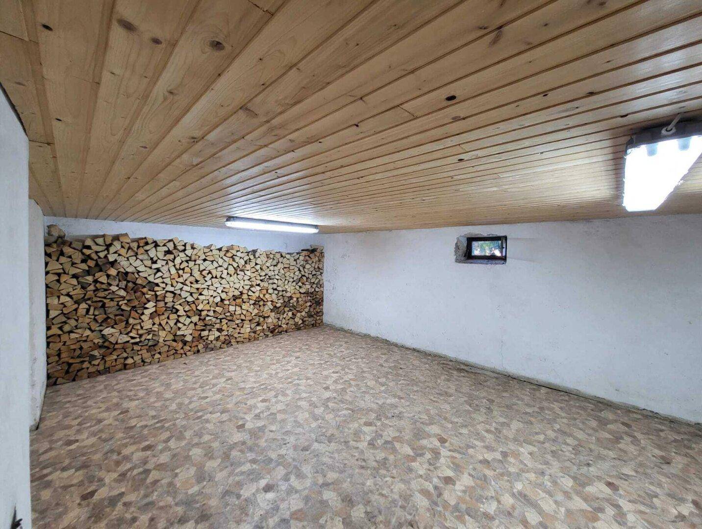 Kellerraum mit Holzdeckenverkleidung und gestapeltem Brennholz für den Ofen.