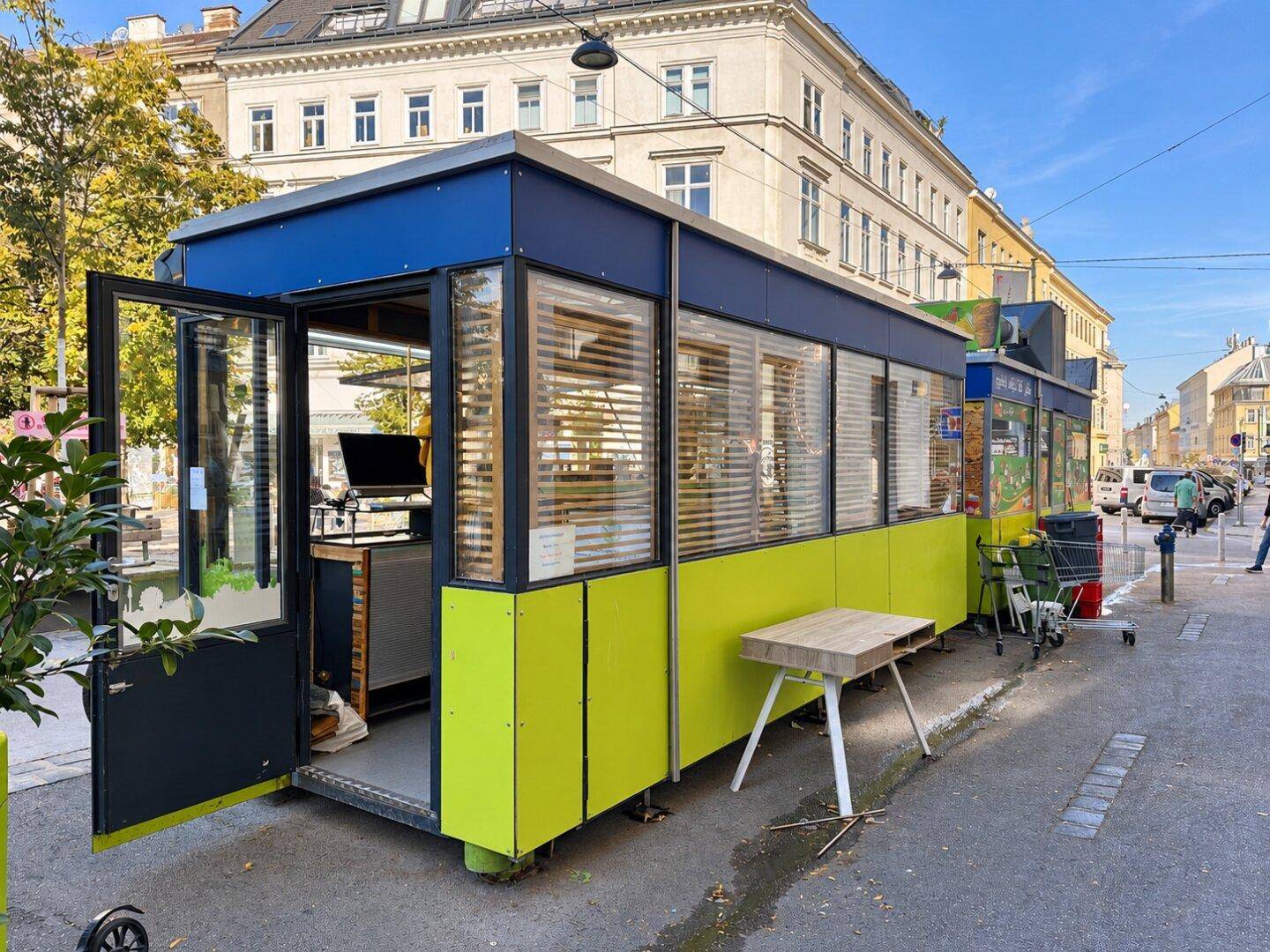 Außenansicht eines modernen Verkaufsstands mit hellgrünen und dunkelblauen Paneelen, gelegen an einer städtischen Straße.