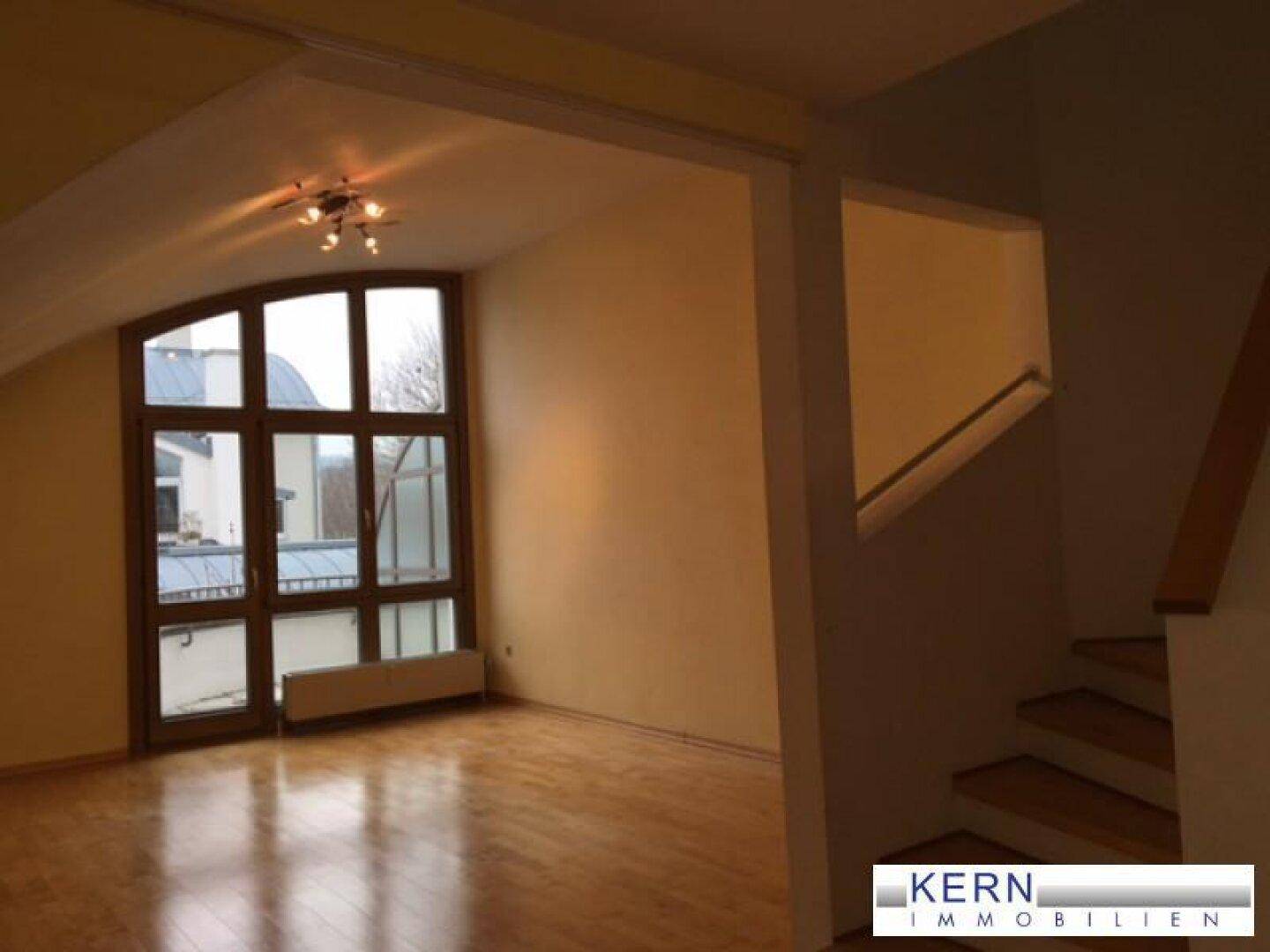 Geräumiges Wohnzimmer mit großen Fenstern, Holzfußboden und Zugang zu einer Treppe.