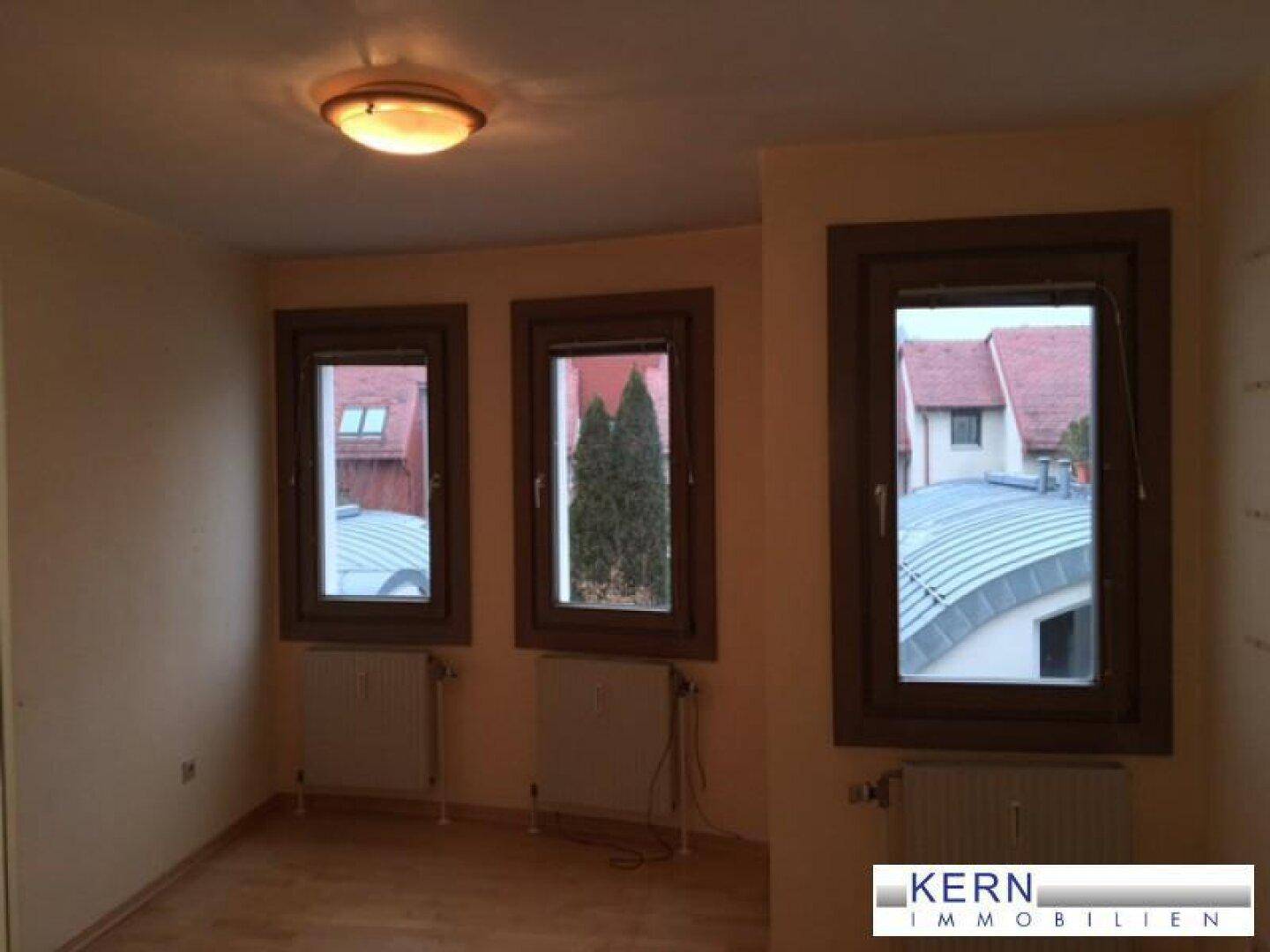 Helles Zimmer mit drei Fenstern und Heizkörpern, bietet einen Blick auf die Dächer der Nachbarschaft.