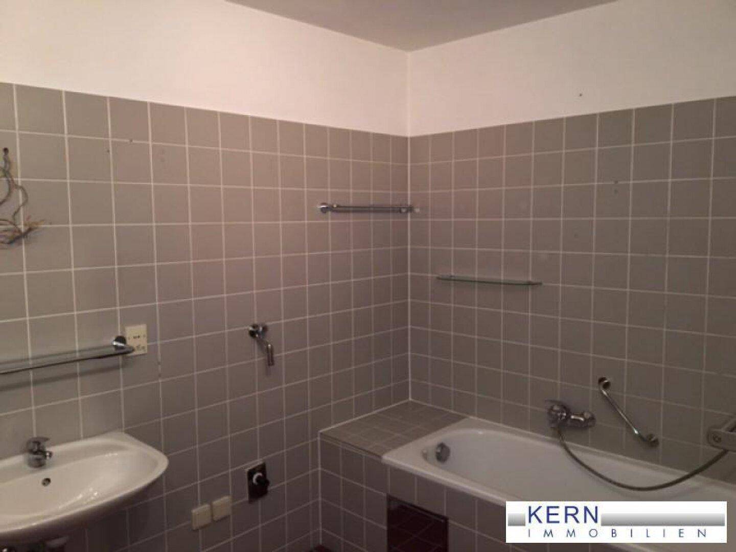 Badezimmer mit Badewanne, Waschbecken und grauen Fliesen, ausgestattet mit Haltegriffen.