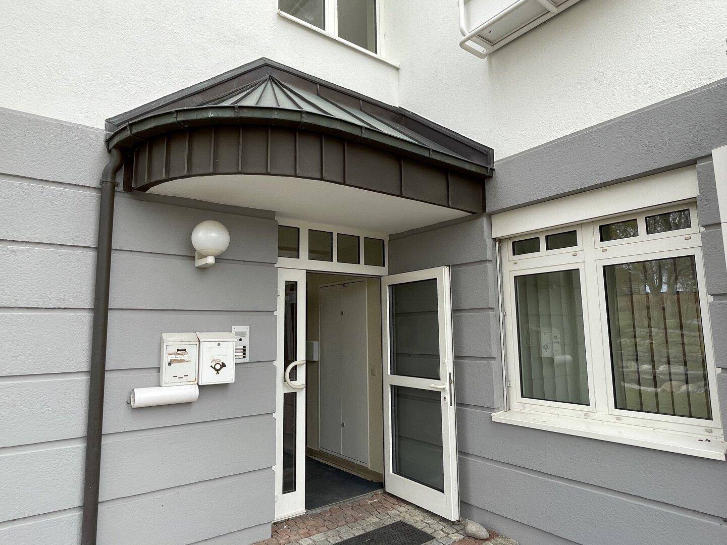 Eingangsbereich eines Gebäudes mit dunklem Vordach, weißer Tür und Briefkästen an der Wand.
