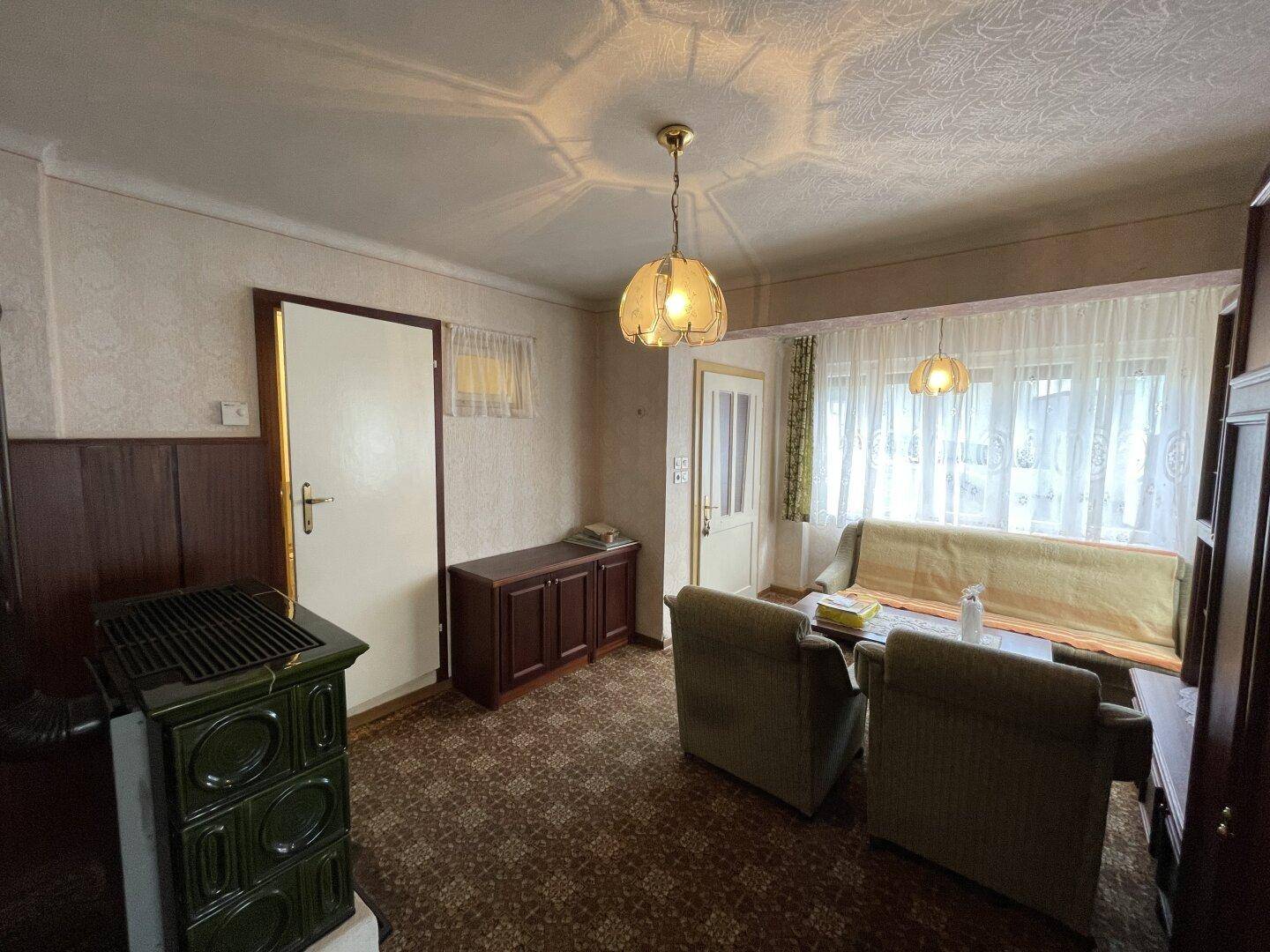 Wohnzimmer mit Teppichboden, traditionellen Möbeln und einem grünen Kachelofen.