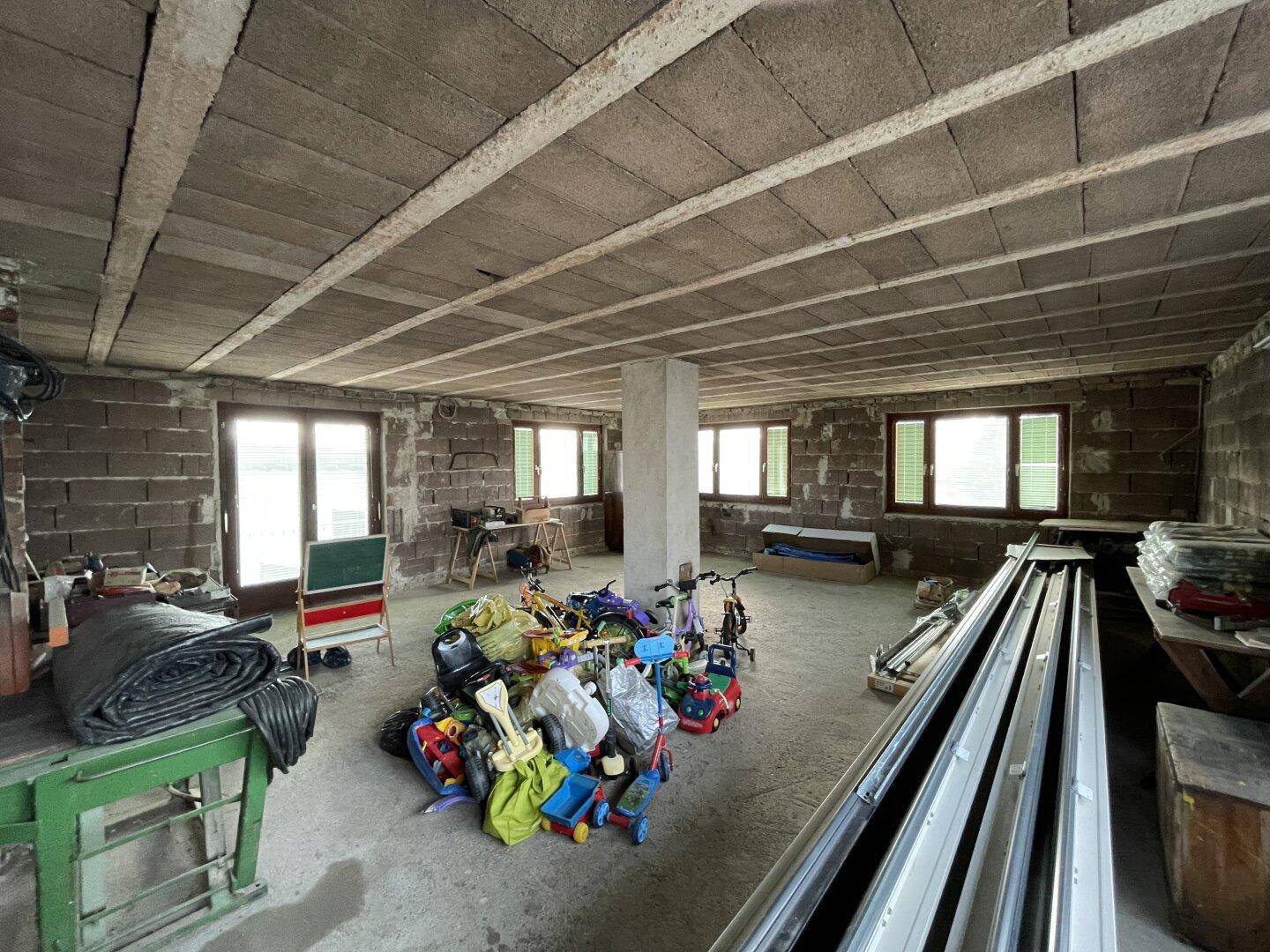Unfertiger Kellerraum mit Betonboden, Fenstern und gelagerten Gegenständen.