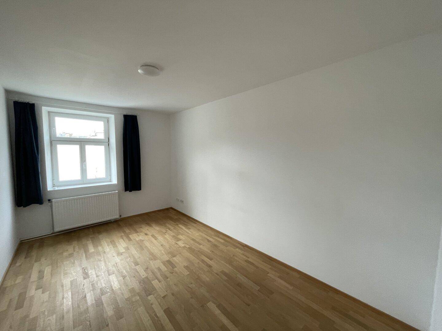 Helles Zimmer mit Parkettboden, weißen Wänden und einem Fenster mit dunklen Vorhängen.