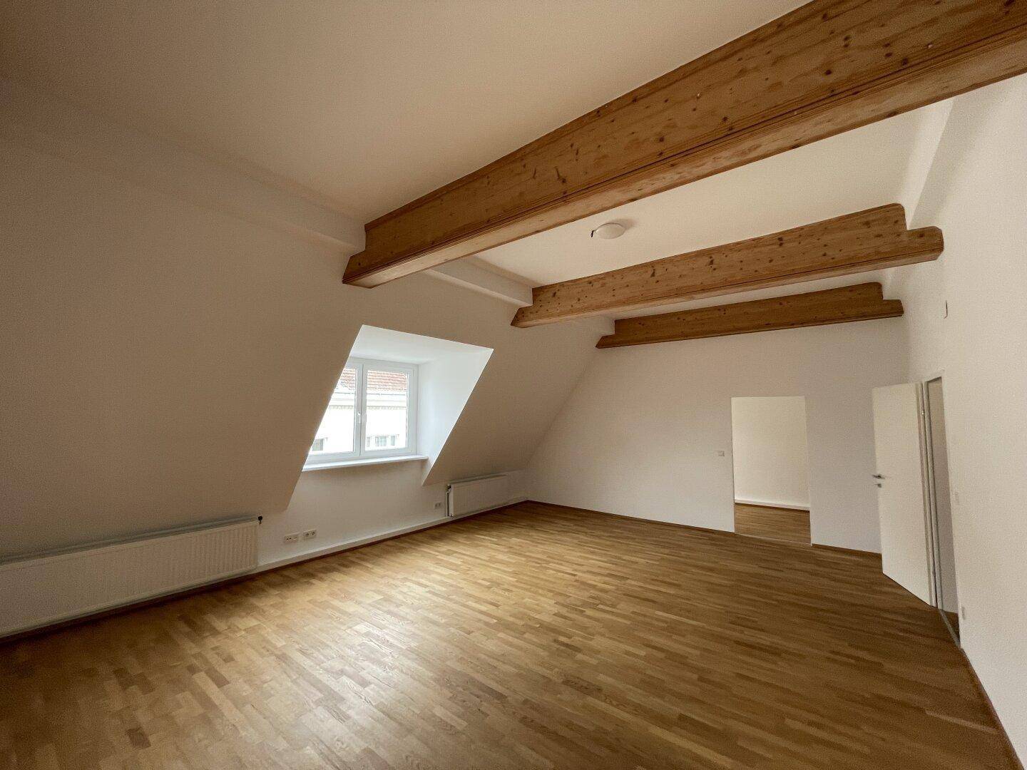 Heller Wohnraum im Dachgeschoss mit schrägen Wänden, Holzbalken und Parkettboden.