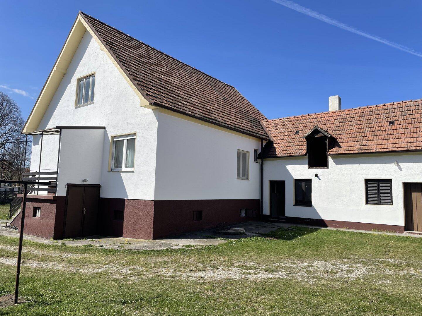 Seitenansicht eines ländlichen Hauses mit weißer Fassade, braunem Sockel und rotem Ziegeldach.