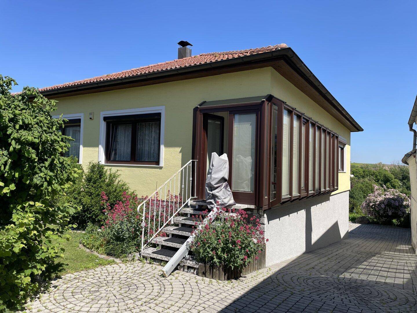 Seitenansicht des Hauses mit Wintergarten, Außentreppe und gepflasterter Zufahrt.