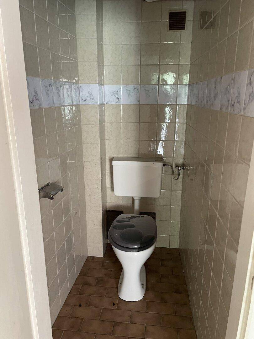 Kleines WC mit beigefarbenen Fliesen, einer Toilette und einem Lüftungsgitter an der Decke.
