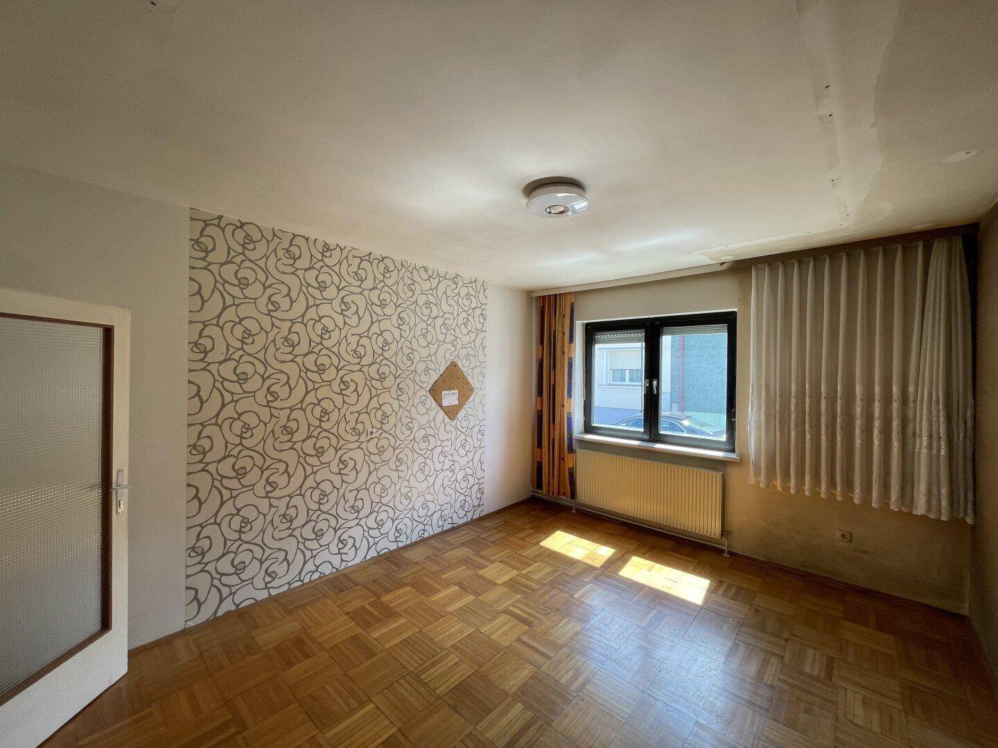 Helles Zimmer mit Parkettboden, gemusterter Tapete und großem Fenster.