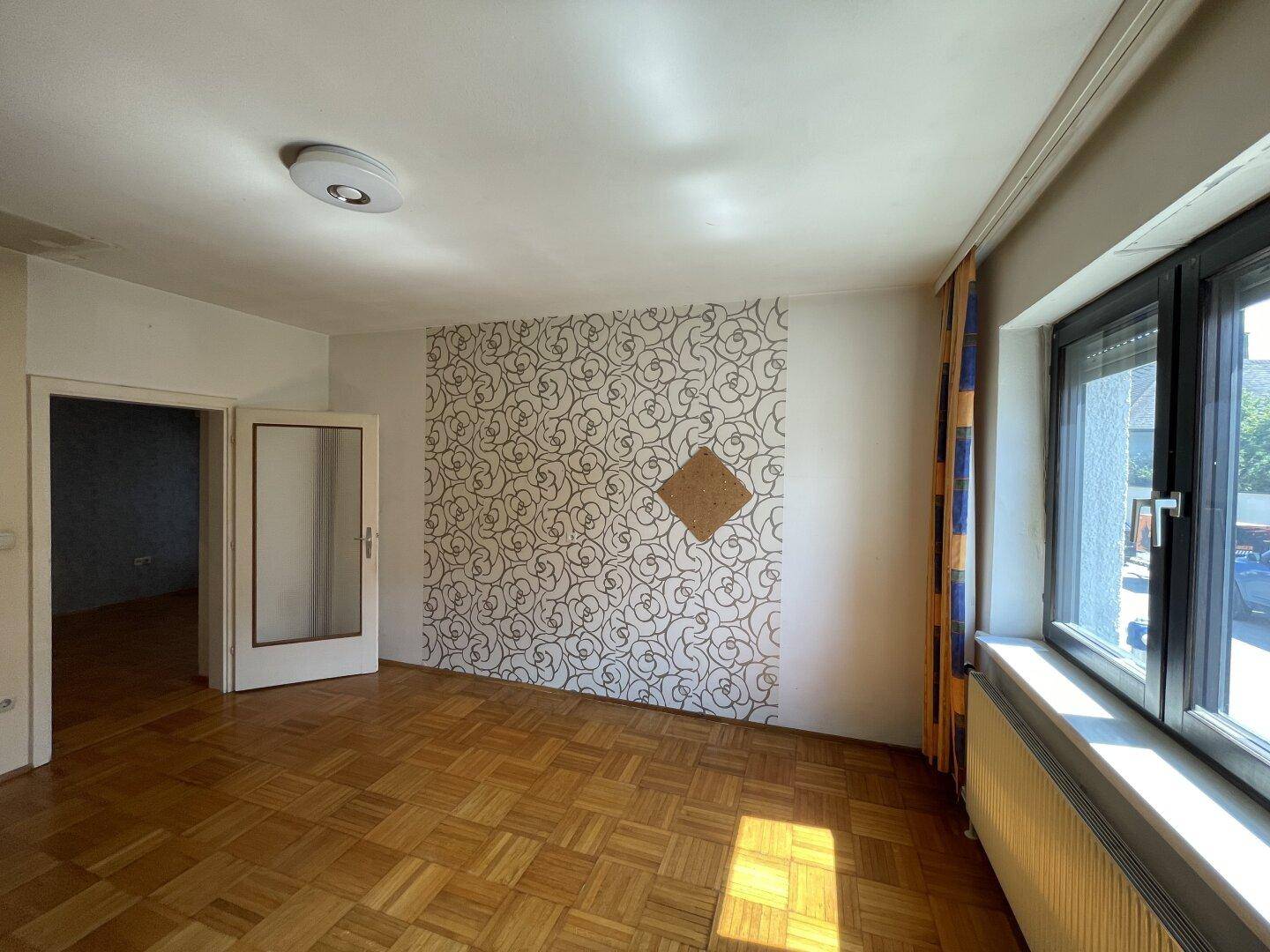 Geräumiges Zimmer mit Parkettboden, Fenster und Zugang zu einem weiteren Raum.