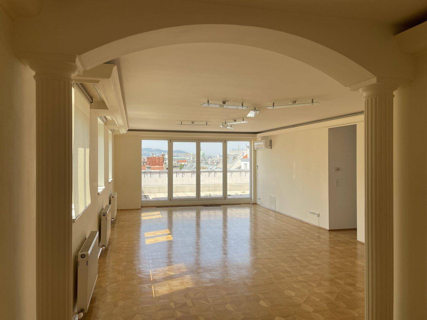 Geräumiges Wohnzimmer mit Parkettboden, großen Fenstern und Blick auf die Stadt.