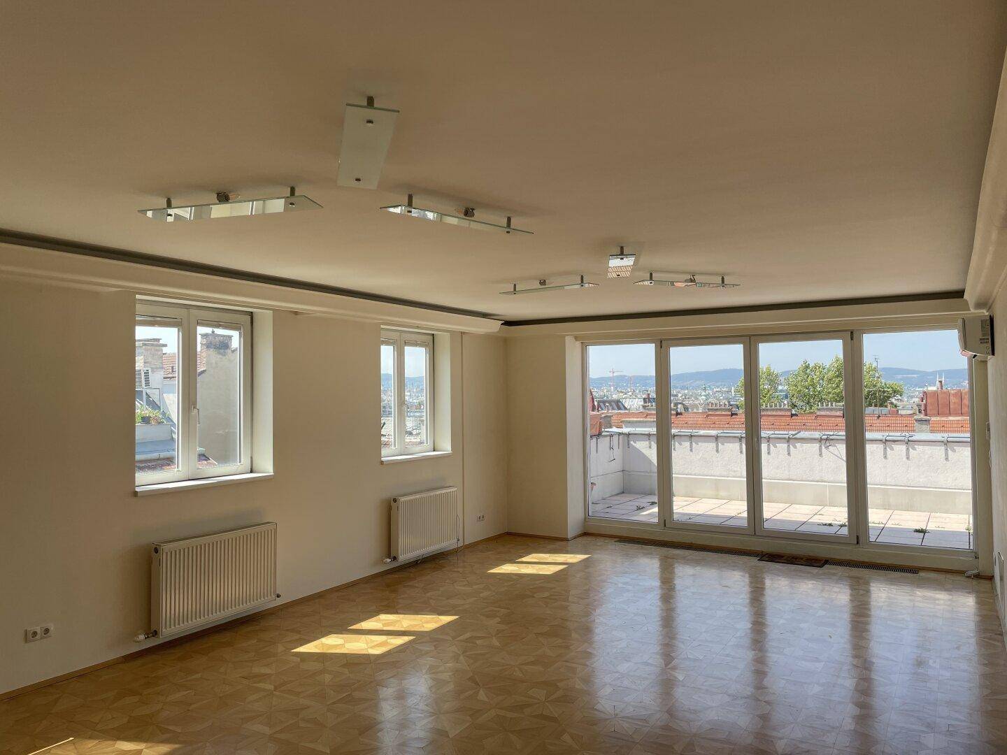 Lichtdurchflutetes Wohnzimmer mit Parkettboden, Heizkörpern und Zugang zur Dachterrasse.