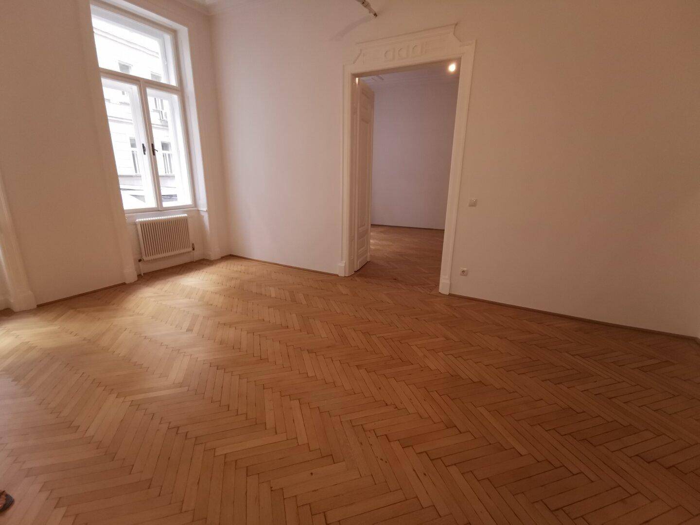 Leerer Raum mit elegantem Fischgrätparkett, hohen Decken und einem Fenster, das den Raum mit Tageslicht füllt.