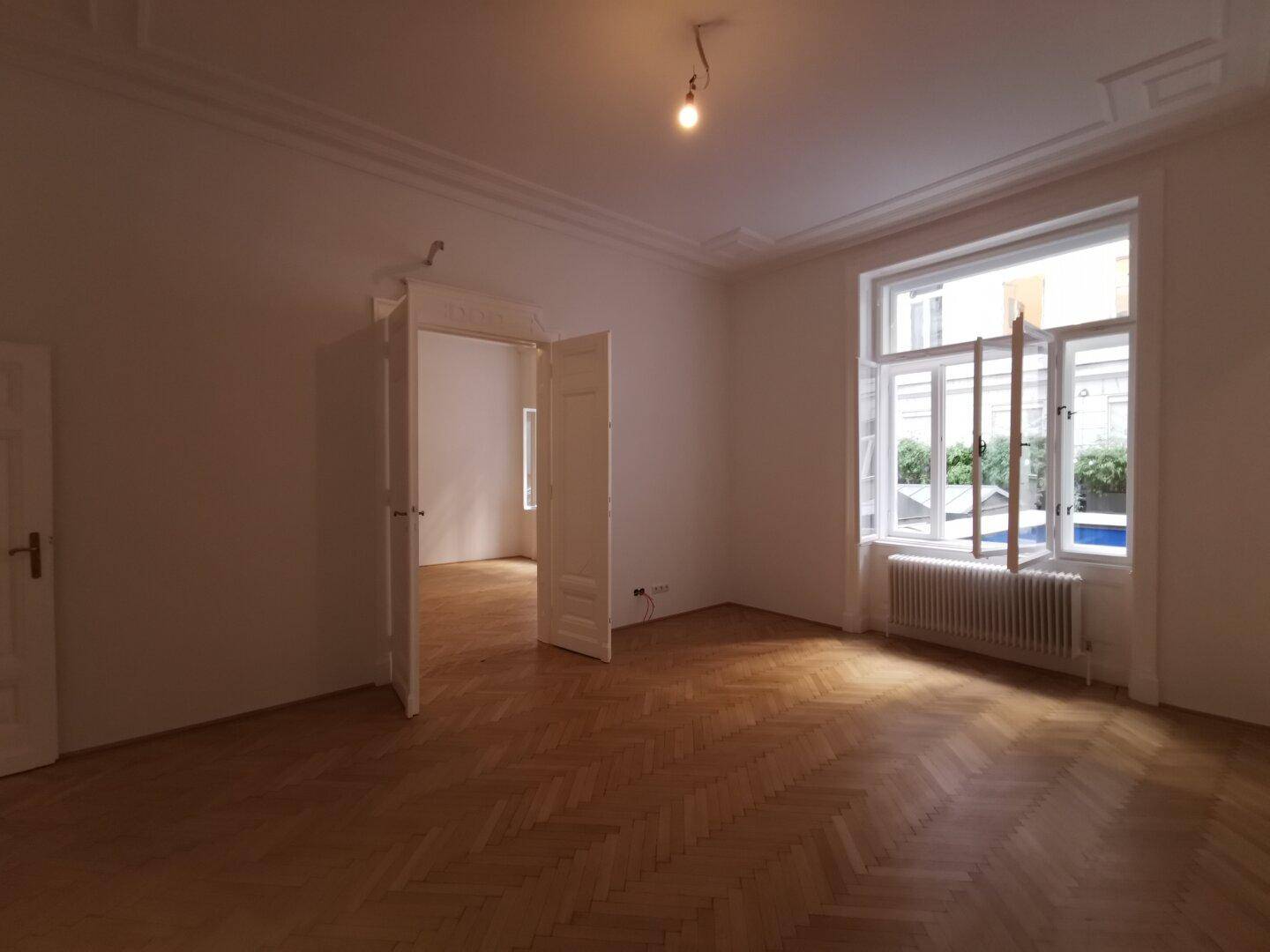 Heller Wohnraum mit Fischgrätparkett, hohen Decken und einem großen Fenster mit Blick auf den Innenhof.