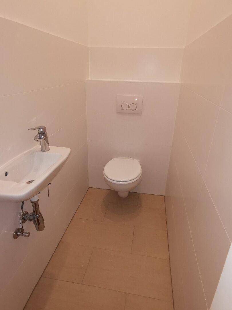 Modernes Gäste-WC mit wandhängender Toilette, kleinem Waschbecken und hellen Fliesen an Wänden und Boden.