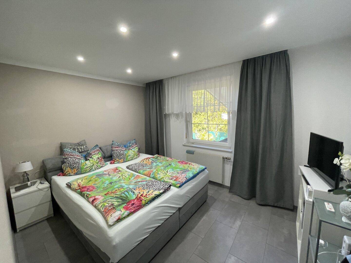 Helles Schlafzimmer mit Doppelbett, farbenfroher Bettwäsche und großem Fenster.