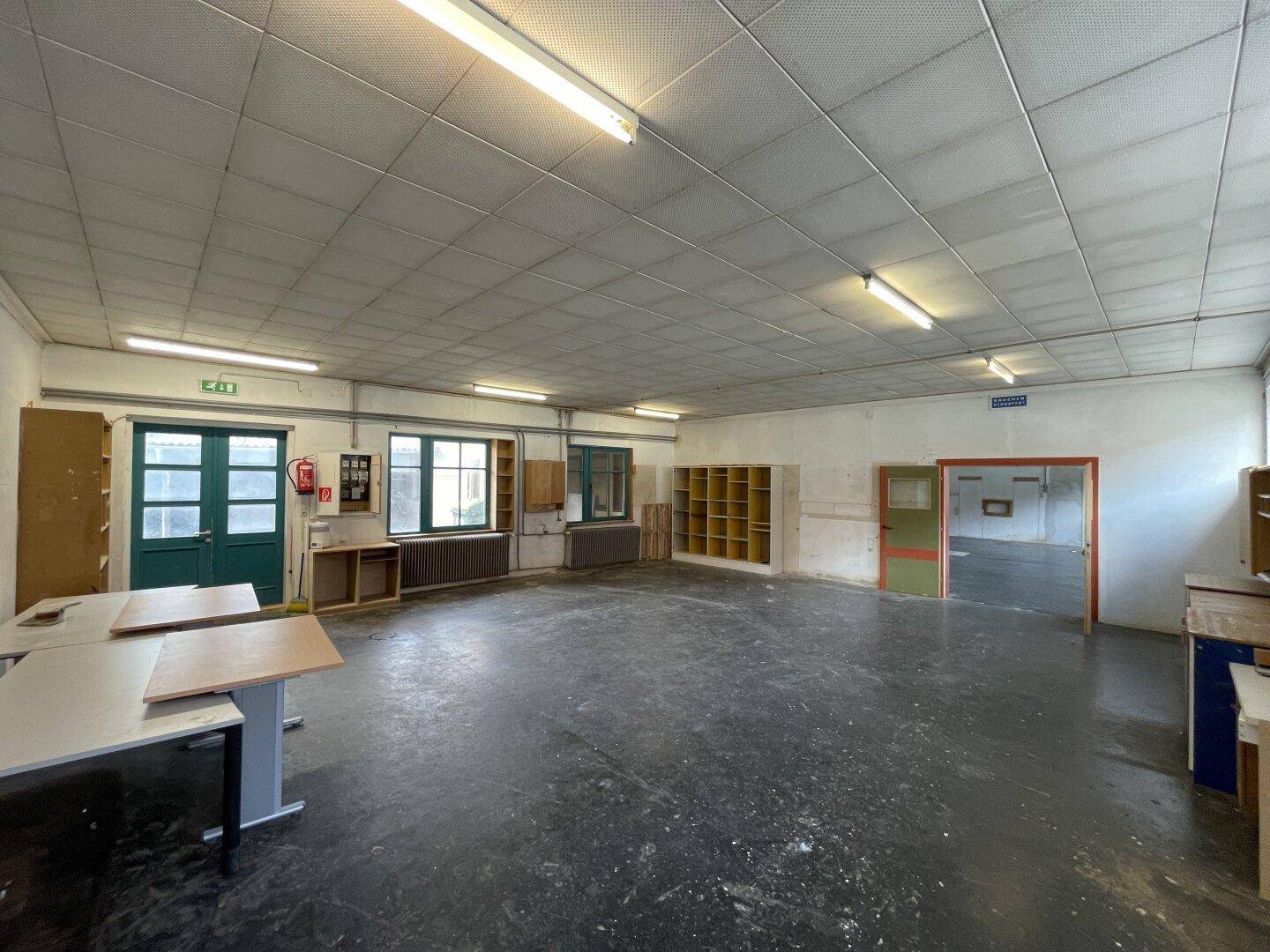 Großer Arbeitsraum mit dunklem Boden, Regalen und mehreren Fenstern, ideal für gewerbliche Nutzung.
