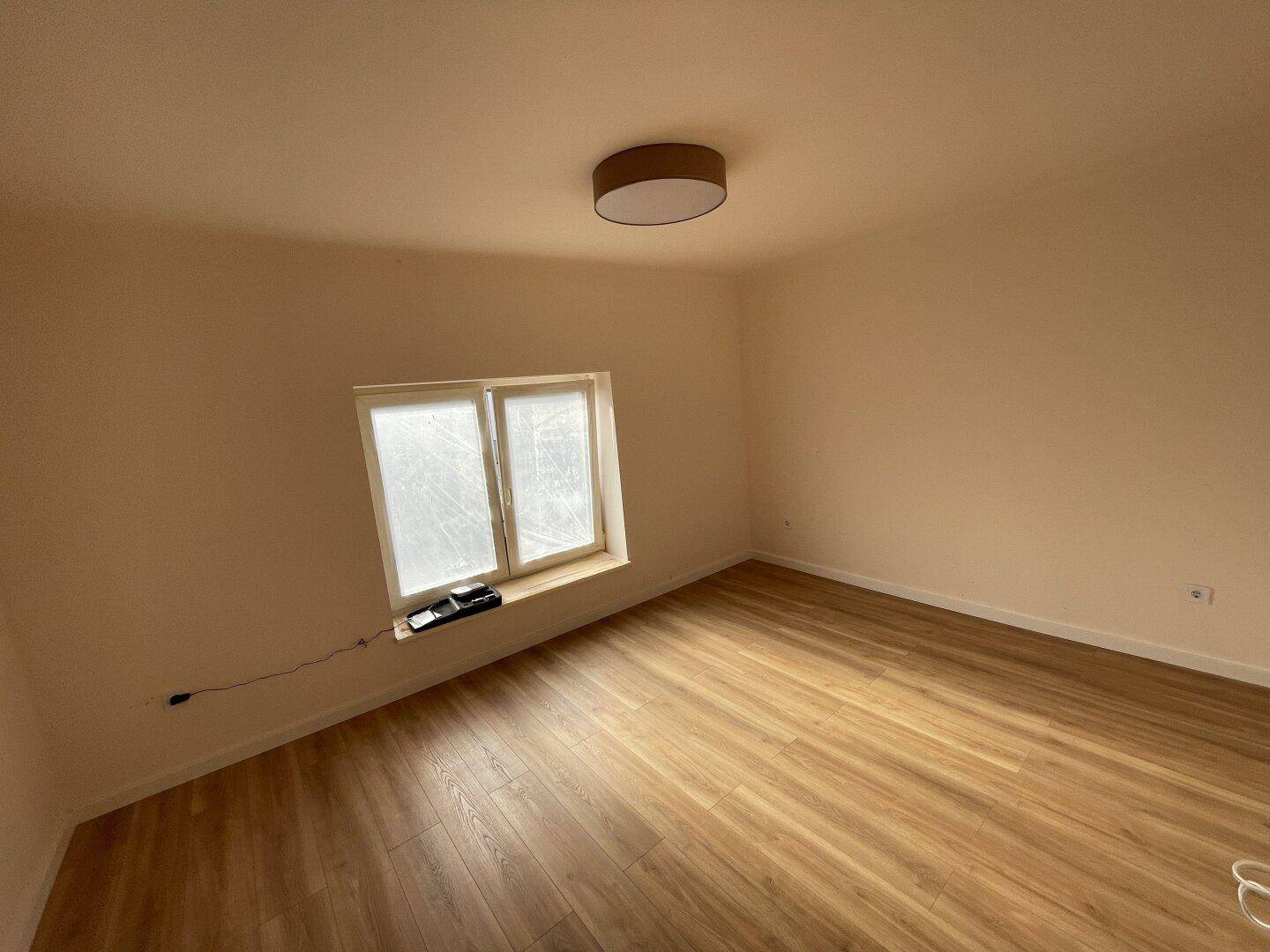 Leeres Zimmer mit hellem Laminatboden und einem Fenster, das Tageslicht hereinlässt.
