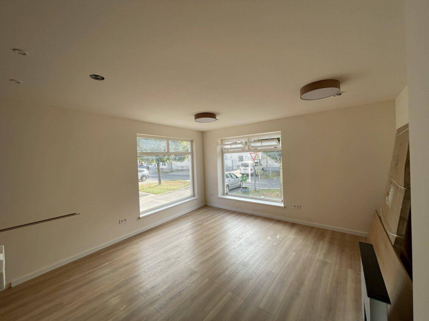 Heller Raum mit Laminatboden und großen Fenstern, die einen Blick auf die Straße und Bäume bieten.