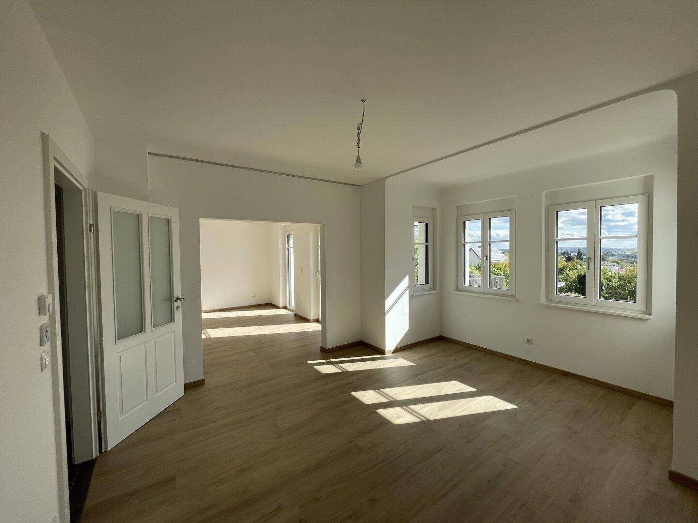 Geräumiges, helles Wohnzimmer mit Holzboden und mehreren Fenstern, die einen schönen Ausblick ermöglichen.