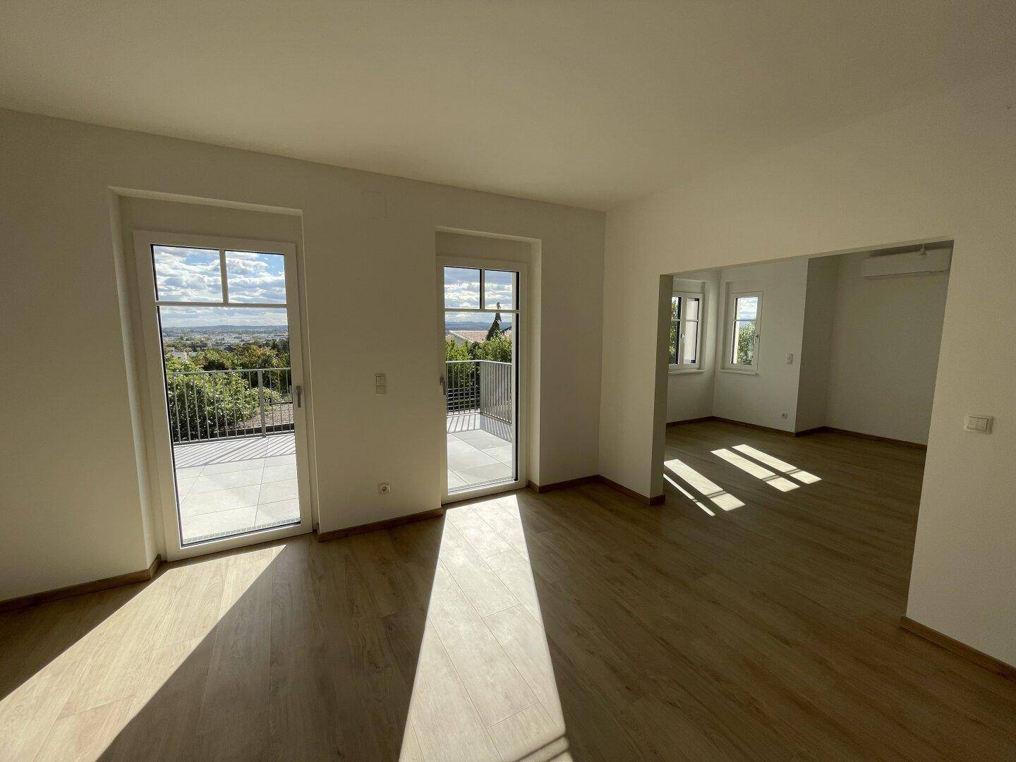 Helles Wohnzimmer mit Holzboden und großen Glastüren, die Zugang zur Terrasse und weitem Ausblick bieten.