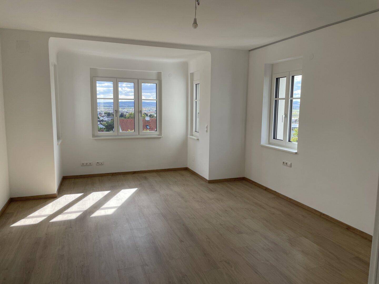 Helles Zimmer mit Holzboden und großen Fenstern, die viel Tageslicht hereinlassen und eine schöne Aussicht bieten.