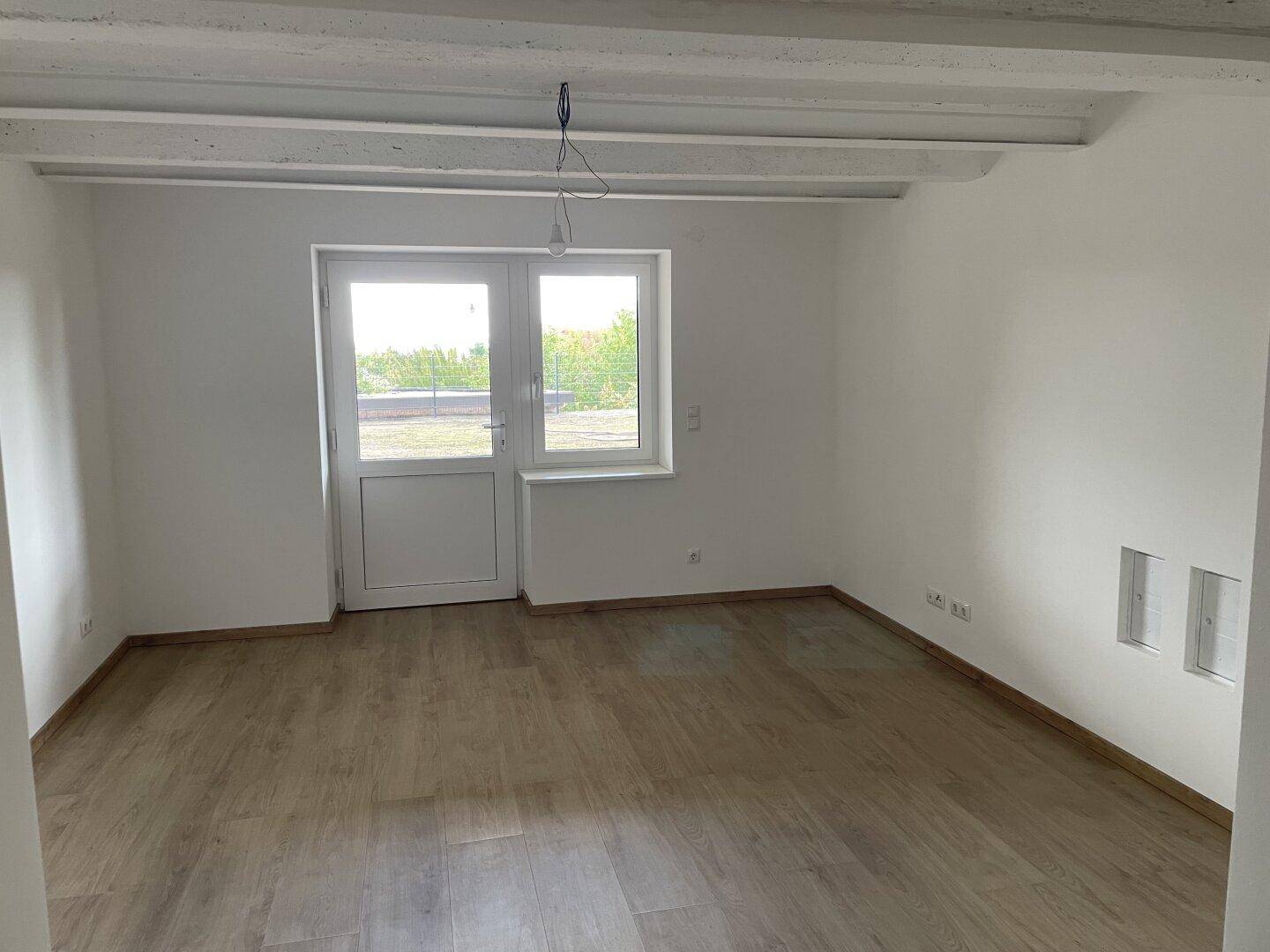 Helles Zimmer mit Holzboden, weißen Wänden und einer Glastür, die zu einem Außenbereich führt, mit sichtbaren Deckenbalken.