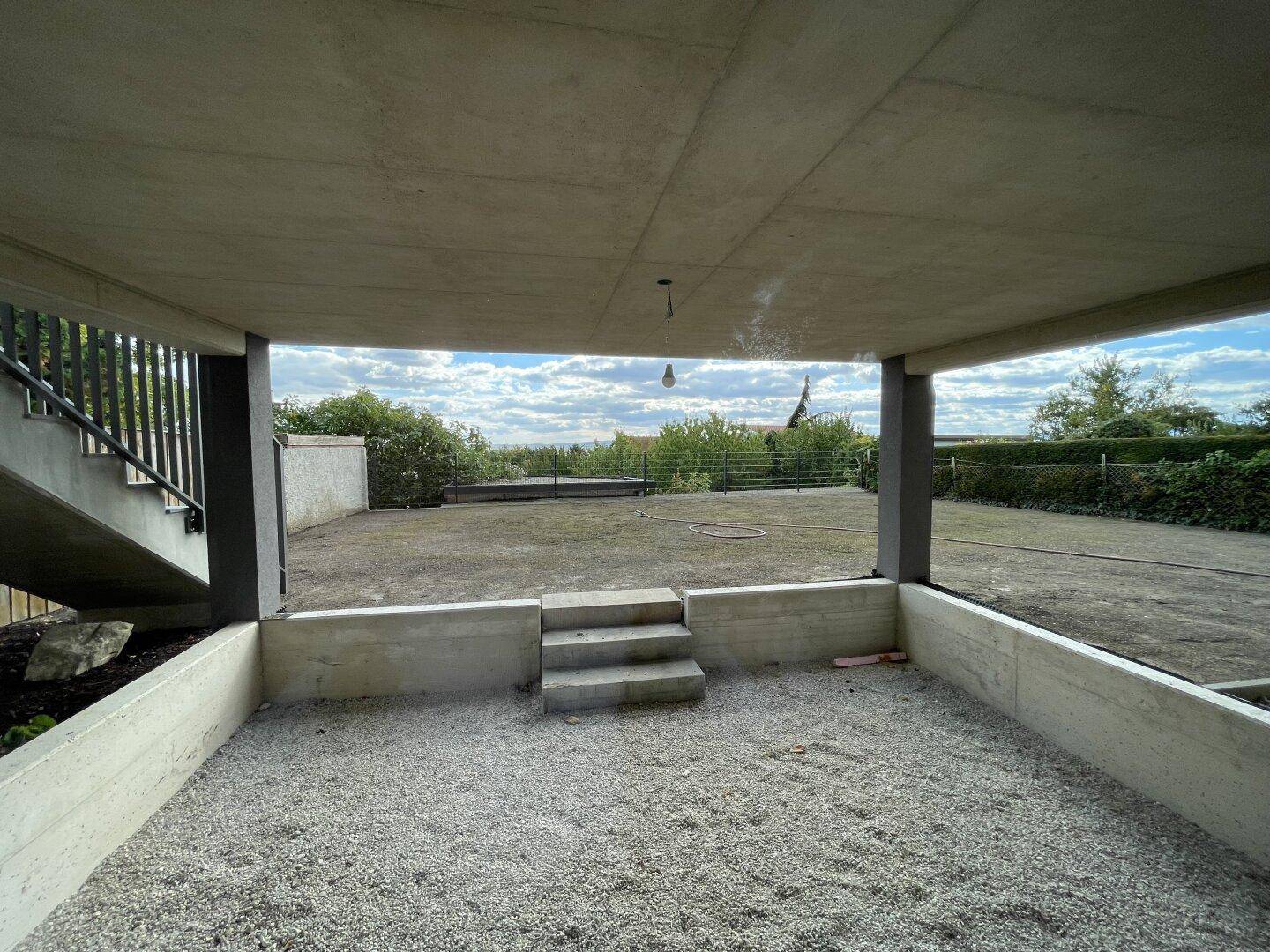 Unfertiger Außenbereich unter einer Betonkonstruktion mit Kiesboden und Blick auf den Garten und die Landschaft.