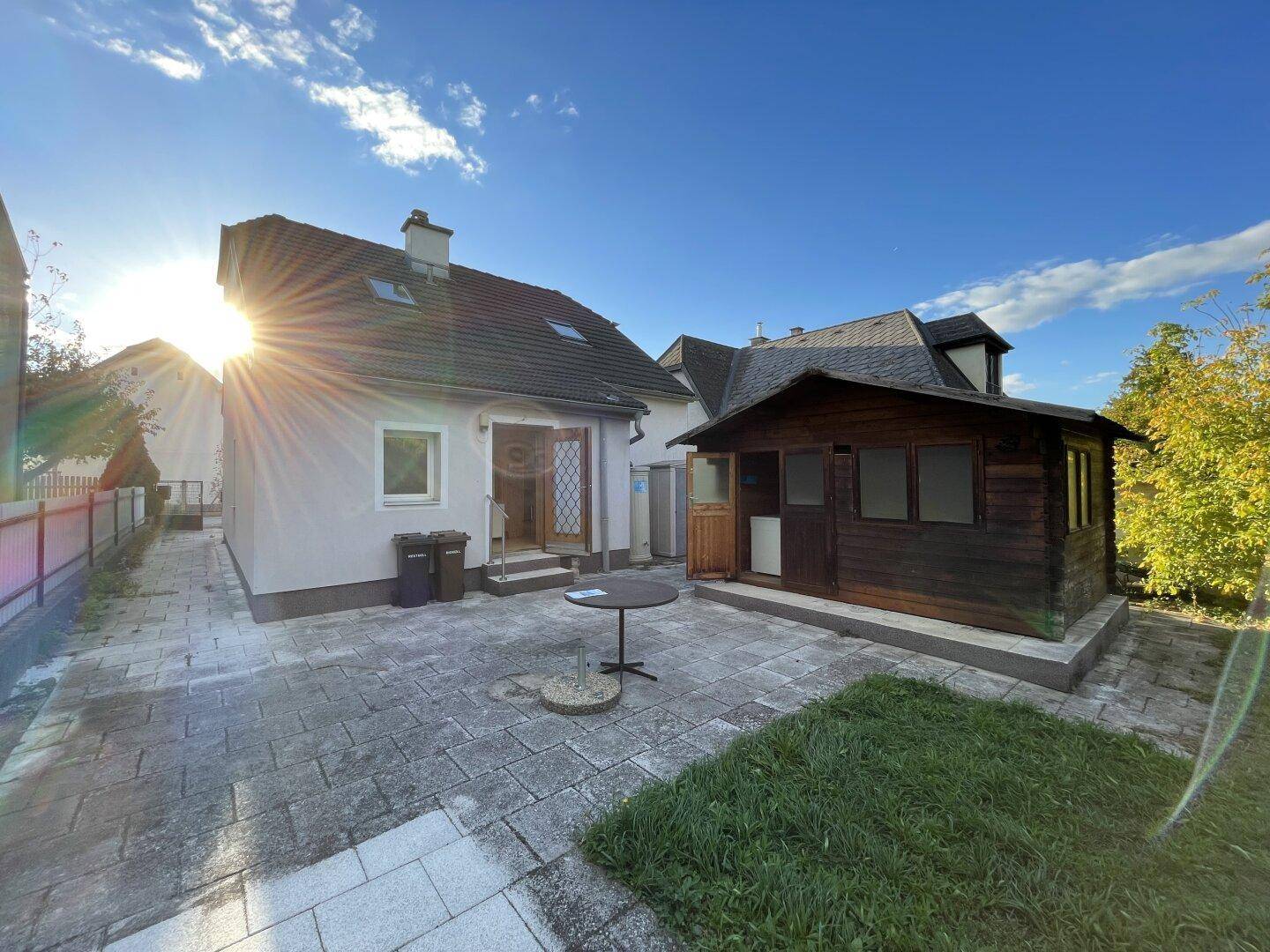 Gepflasterte Terrasse mit kleinem Gartentisch und Holzschuppen im sonnigen Gartenbereich.