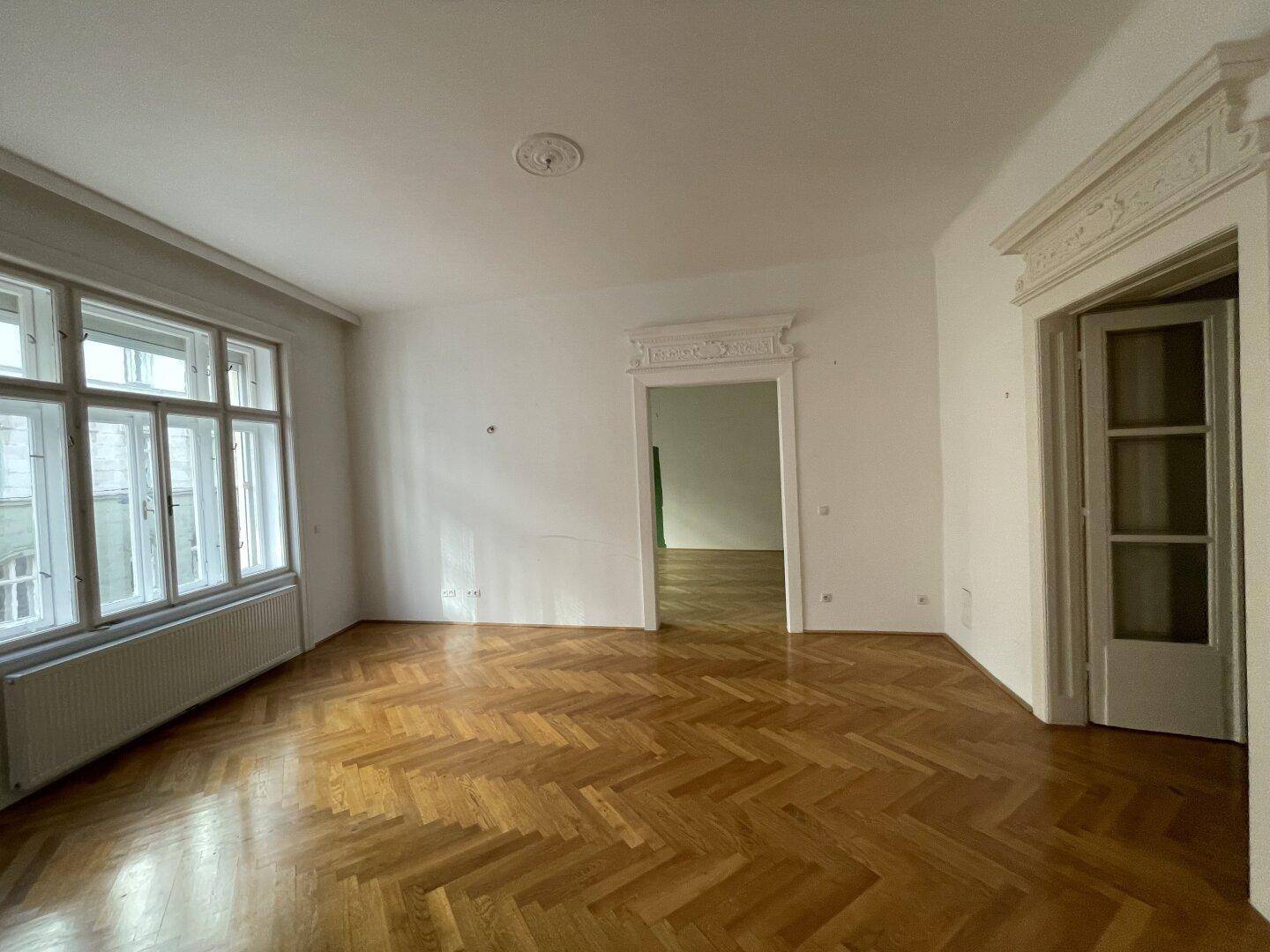 Leeres Wohnzimmer mit Fischgrätparkett, hohen Decken und Blick auf die Straße.